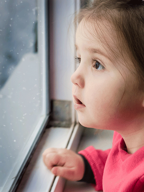 Mädchen schaut aus einem Fenster. Vor dem Fenster fällt Schnee herab.